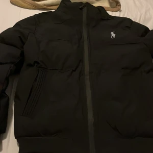 Svart pufferjacka från Polo Ralph Lauren - Svart pufferjacka från Polo Ralph Lauren med klassisk logga broderad på bröstet. Jackan har hög krage, dragkedja framtill och ribbade muddar vid ärmslut och midja. Fodrad insida i blankt material och två snedställda fickor framtill.