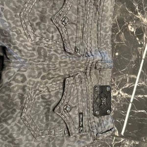 Grå leopardmönstrade Miss Me cargo - Sitter som S/M. Snygga grå cargo-byxor från Miss Me med leopardmönster och coola detaljer som dekorativa knappar och broderad logga på bakfickan. Byxorna har raka ben och kontrastsömmar som ger extra edge. Perfekt för dig som vill sticka ut med din stil.