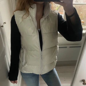 Beige puffer väst med ribbad krage - Snygg beige puffer väst med dragkedja framtill. Västen har en ribbad, ljusgrå krage och två fickor med dragkedja. Justerbara band både i midjan och armarna. 