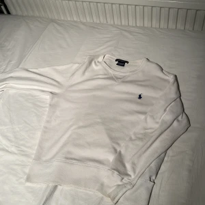 Vit sweatshirt från Ralph Lauren Sport - Vit långärmad sweatshirt från Ralph Lauren Sport med klassisk blå logga broderad på bröstet. Tröjan har rund halsringning och är tillverkad i en mjuk bomulls- och polyestermix. Enkel och clean stil som funkar till allt. Ett litet hål längst ner på vänster sida men syns inte när man har på sig den.