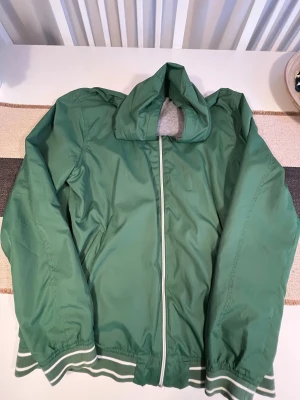 Grön vindjacka från Livergy - Snygg grön vindjacka från Livergy med huva och vita detaljer på dragkedjan samt ribbade muddar med vita ränder. Insidan är fodrad med grått tyg och har en innerficka. Jackan är tillverkad i polyester och bomull, perfekt för dig som gillar sportig stil. Unisex.