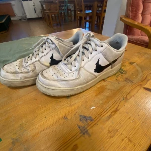 Nike Air Force 1 med orange detalj - Nike Air Force 1 sneakers i vitt skinn med orange häl och svart, edgy swoosh på sidan. Klassisk låg modell med snörning och perforerad tå för extra ventilation. Platt sula och rund tå, perfekt för dig som gillar streetstyle och unika detaljer. Några hål på insidan.