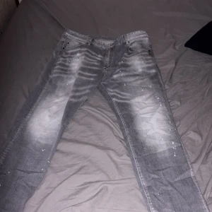 Grå jeans från Dsquared2 med splatter - Säljer ett par grå jeans från Dsquared2 i storlek 38. De har coola slitningar och vita färgstänk över hela byxorna, samt en röd patch med texten 'Caten Heated' bak. Jeansen har klassisk femficksdesign och är tillverkade i bomull.