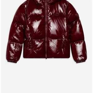 Vinröd pufferjacka från Tommy Hilfiger - Snygg vinröd pufferjacka från Tommy Hilfiger med glansig finish och huva. Jackan har dragkedja framtill, två stora fickor och diskret logga på bröstet. Perfekt för dig som vill ha en varm och trendig look under kalla dagar. Finns inga tecken på användning.