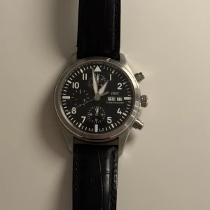 IWC Pilot's Chronograph svart - Snygg IWC Pilot's Chronograph med svart urtavla, vita siffror och detaljer, samt dag- och datumvisning. Boetten är i silverfärgat stål och armbandet är svart med läderlook. Klockan har tre subdials och klassisk rund form med robusta tryckknappar på sidan.