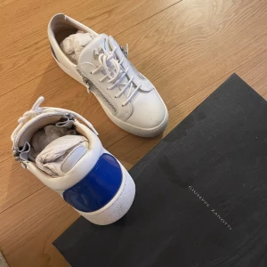 Giuseppe Zanotti vita sneakers med blå detalj - Säljer ett par vita sneakers från Giuseppe Zanotti med blå hälpanel, snörning och silverfärgade dragkedjor på sidorna. Skorna har en platt sula och är tillverkade i skinn med metallisk logga på plösen. Riktigt snygga och unika detaljer som sticker ut.