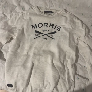 Vit stickad tröja från Morris R Club - Snygg vit stickad tröja från Morris R Club med svart tryck framtill och ribbade muddar vid hals, ärmslut och nederkant. Klassisk rund hals och långärmad modell, perfekt för dig som gillar stilrena och sportiga vibes. Jätte snygg men passar ej mig. Texten är läder.