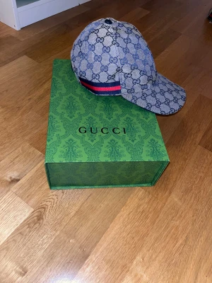 Gucci keps med blått GG-mönster - Säljer en snygg Gucci keps i storlek M med klassiskt blått GG-mönster och en ikonisk röd-blå rand på sidan. Kepsen är tillverkad i manchester och har justerbar rem baktill. Kommer med original dustbag och grön Gucci-box.