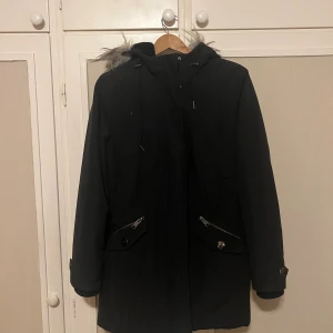 Svart vinterjacka - Säljer denna kättemysiga och praktiska vinterjackan då den inte kommer till användning! Använd men i fint skick med praktiska fickor, fluffig insida, fluffig luva och ett band på vardera sida som man kan använda för att dra åt midjan! Stängs med dragkedja och tryckknappar framtill.