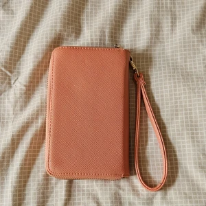 Rosa plånbok med handledsrem - Snygg rosa plånbok med praktisk handledsrem och guldfärgade dragkedjor. Plånboken har flera fack för kort och sedlar, samt ett extra fack med dragkedja på utsidan. 