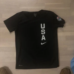 Svart Nike USA t-shirt Dri-Fit - Svart t-shirt från Nike med Dri-Fit material och trycket 'USA' i vitt på framsidan. Kortärmad modell med Nike-logga och patch på ena ärmen. Perfekt för dig som gillar sportig stil och vill representera USA.