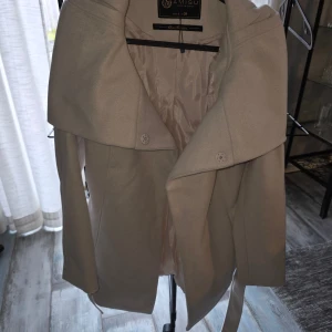 Beige kappa från Amisu med bälte, oanvänd  - Snygg beige kappa från Amisu med bred krage och bälte i midjan. Jackan har en klassisk design med knappar framtill och slät yta. Perfekt för dig som gillar stilrena och tidlösa plagg.