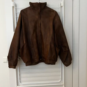 Brun oversized skinnjacka med hög krage - Säljer en brun vintage skinnjacka med hög krage och elastisk midja. Jackan har två fickor framtill. Insidan har ett vitt tyg med svarta ränder. Perfekt för dig som gillar retrovibbar 