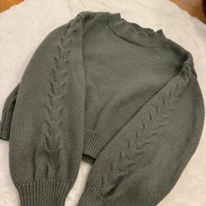 Grå stickad tröja med flätmönster - Snygg grå stickad tröja med flätade detaljer längs ärmarna och ribbad kant vid hals, ärmslut och nederkant. Tröjan har en lite högre hals med vågig kant och är långärmad. Perfekt för dig som gillar mysiga och trendiga plagg.