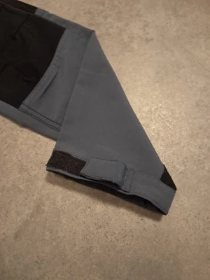 Grå och svarta cargopants från Strova - Säljer ett par grå cargopants från Strova med svarta förstärkningar på knän och bakdel. Byxorna har flera praktiska fickor med dragkedja och knappstängning, samt justerbar midja.. Perfekt för dig som gillar funktionella och snygga byxor.