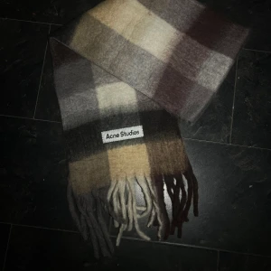 Rutig halsduk Acne Studios  - Snygg rutig halsduk från Acne Studios . Färgerna går i beige, brun, grå och vit med mörka och ljusa toner. Klassisk design med fransar i ändarna och tydlig Acne Studios-label. Perfekt för att lyfta din vinterstil.