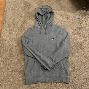 Grå hoodie från Lyle & Scott M - Säljer en grå hoodie från Lyle & Scott med klassisk gul logga på bröstet. Tröjan har huva med snörning och en stor magficka framtill. Perfekt för chill dagar och enkel att matcha med jeans eller joggers. Den är i superfint skick och har inte använts så mycket. Hör av er om ni har frågor!