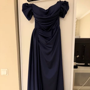 Midnattsblå långklänning med puffärm - Säljer en elegant oanvänd marinblå långklänning med offshoulder puffärmar och draperad överdel. Klänningen har en snygg slits och dragkedja i ryggen. Materialet är slätt och har ett fint fall, perfekt för bal eller fest. Stilren och modern look som verkligen sticker ut. De står att de är storlek 38, men passar mig som är storlek 36 och S/XS prefekt. 