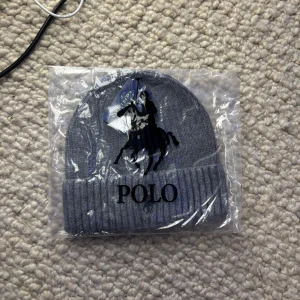 Grå stickad mössa från Polo Ralph Lauren - Snygg grå mössa från Polo Ralph Lauren med klassisk ribbad kant och broderad svart logga framtill. Mössan är stickad och har en enkel, stilren form som passar perfekt för kalla dagar. Materialet är mjukt och håller dig varm hela vintern.