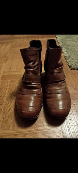 Bruna fodrade boots från Rieker - Säljer ett par bruna boots från Rieker i mjukt skinn med rund tå och dekorativa sömmar. Skorna har dragkedja på sidan och ett snyggt spänne med metallknapp. Insidan är fodrad med vitt, fluffigt material som håller fötterna varma under kalla dagar. Det är ett litet skavmärke på ena som jag tagit bild på, i övrigt gott skick. 