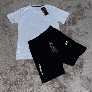 Vit Under Armour t-shirt med holografisk tryck - Snygg vit t-shirt från Under Armour med holografiska detaljer och logga på bröstet, axeln och nederkanten. T-shirten har en klassisk rund hals och korta ärmar, perfekt för träning eller chill. Materialet är lätt och andas, vilket gör den skön att bära.