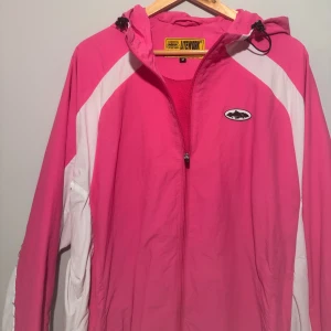 Rosa och vit vindjacka Corteiz  - Säljer en snygg vindjacka från Corteiz i rosa och vitt med huva och dragkedja framtill. Jackan har coola detaljer som logopatch på bröstet och broderad text på ryggen. Perfekt för dig som vill sticka ut med färg och streetstyle. 