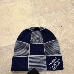 Louis Vuitton rutig mössa i blått - Snygg mössa från Louis Vuitton med rutigt mönster i marinblått och grått. Tillverkad i lyxig mix av kashmir, ull och silke. Broderad text 'Marque L. Vuitton Deposee' framtill. Perfekt accessoar för dig som vill sticka ut med stil.