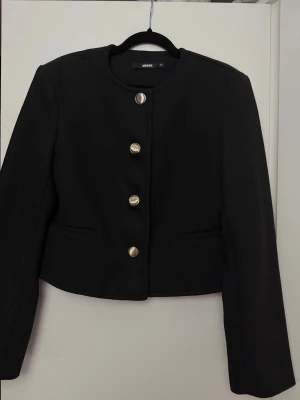 Svart kavaj med guldknappar Bik Bok - Stilren svart kavaj från Bik Bok i storlek XS med rund halsringning och fem snygga guldknappar framtill. Jackan är croppad och har två fickor fram. Perfekt för dig som gillar klassisk och trendig stil.
