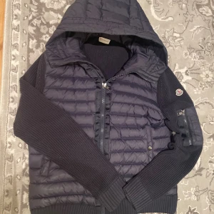 Moncler Cardigan - Snygg mörkblå Cardigan från Moncler med stickade ärmar och huva. Jackan har quiltad front, dragkedja och Moncler-logga på ärmen. Materialmix med dunfyllning och stickad polyester ger både värme och stil. Perfekt för dig som vill ha en cool look i höst och vinter.