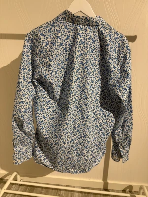 Blommig vit och blå skjorta Selected - Snygg skjorta från Selected med blommigt mönster i blått på vit botten. Skjortan har klassisk krage, knappar framtill och långa ärmar. Tillverkad i mjuk bomull som känns skön mot huden. Perfekt för dig som gillar att sticka ut med stilrena detaljer.