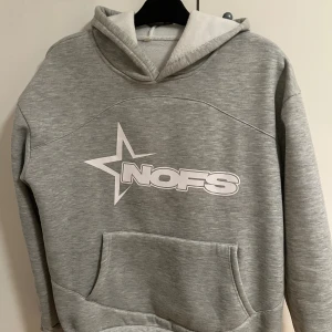 Grå hoodie från NOFS med tryck - Grå hoodie från NOFS med stor vit stjärn- och logotyptryck på bröstet. Tröjan har huva, magficka och långa ärmar. Mjuk och skön, perfekt för chill dagar. Enkel design med en modern touch som passar till jeans eller joggers.