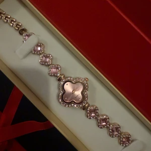 Roséguld klocka med rosa stenar - Säljer en elegant klocka i roséguld med fyrklöverformad urtavla och gnistrande stenar runt både urtavlan och länkarna. Länken har rosa fyrkantiga stenar med glittrande infattning. Perfekt accessoar för dig som gillar bling och unik design.
