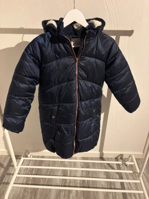 Mörkblå pufferjacka från Lindex - Säljer en mörkblå pufferjacka från Lindex med huva och mysigt ljust foder. Jackan har långa ärmar, två fickor med knapp och en snygg dragkedja i kopparfärg. Perfekt för kalla dagar och har en cool quiltad look.