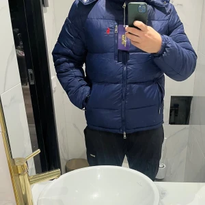 Mörkblå pufferjacka från Polo Ralph Lauren - Snygg mörkblå pufferjacka från Polo Ralph Lauren med röd broderad logga på bröstet. Jackan har huva, flera dragkedjefickor och quiltad design. Perfekt för kalla dagar och har en modern, loose passform.