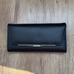 Svart plånbok Classic & Modern - Stilren svart plånbok i slät skinnimitation med guldfärgad detalj framtill. Plånboken har flera kortfack och ett fönster för ID eller foto. Perfekt för dig som vill ha en klassisk och snygg accessoar med plats för allt du behöver.