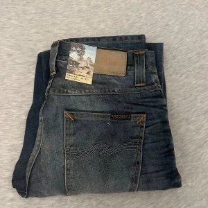 Nudie Jeans Grim Tim blå jeans - Snygga Nudie Jeans Grim Tim i tvättad blå denim med coola slitningar och kontrastsömmar. Klassisk femficksmodell med rak passform och orange detaljer. Märkespatch baktill och unika inskriptioner på insidan. Perfekt för dig som gillar street och casual.