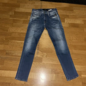 Replay Anbass jeans blå, storlek 27/30 - Snygga Replay Anbass jeans i klassisk blå tvätt med lätt slitningar och raka ben. Modellen har fem fickor, detaljerade sömmar på bakfickorna och normal passform. Perfekt för dig som gillar stilrena och bekväma jeans med modern look.