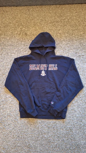 Marinblå 'Belmont Hill' hoodie från Champion - Skön marinblå Belmont Hill hoodie från Champion i storlek M. Använd fåtal gånger och skicket är  i princip som ny, inga slitningar eller defekter. Den har dock lite creasningar, men det går enkelt bort med lite strykning. Passar riktigt bra när du vill ha en lite mer lugnare dag. Skick: 8/10