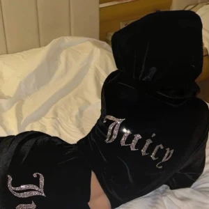 Juicy Couture svart velour hoodie & byxor  - Svart hoodie & byxor från Juicy Couture i mjuk velour med glittrande Juicy-text i strass på ryggen. Tröjan har huva, lång ärm och en loose passform som ger en chill vibe. Perfekt för dig som gillar ikoniska 00-talsstilen och vill sticka ut.