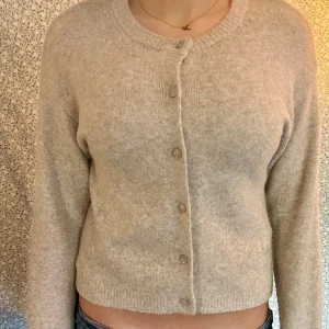 Beige kofta med knappar - Mysig beige kofta med rund hals och knäppning framtill. Tröjan är croppad och har långa ärmar samt ribbade muddar vid hals och nederkant.