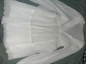 Vit kortklänning med volanger från Nelly - Supersöt vit kortklänning från Nelly med plisserat tyg och volangdetaljer. Klänningen har långärmade ärmar och v-ringad hals. Perfekt för dig som gillar en feminin och fräsch look. Materialet känns lätt och luftigt, vilket gör den skön att bära under varma dagar.