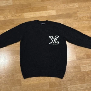 Svart stickad tröja Louis Vuitton - Svart stickad tröja från Louis Vuitton med stort vitt LV-logo på bröstet. Tröjan har rund hals och långa ärmar, perfekt för dig som gillar exklusiva märken och vill ha en clean look med tydlig branding. Materialet är mjukt och ger en skön känsla.