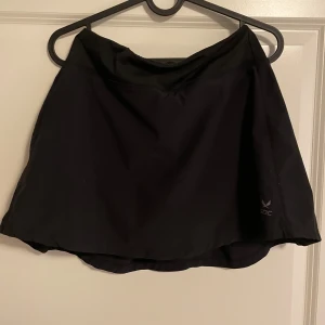 Svart tenniskjol från SOC XS - Sportig svart kortkjol från SOC i storlek XS, perfekt för träning. Kjolen har inbyggda shorts under och är gjord i ett lätt syntetmaterial med teknisk Airflow-funktion som håller dig torr och bekväm. Enkel design utan mönster, med elastisk midja för skön passform. Perfekt om man vill ha kort kjol med extra bekvämligheten av shorts.