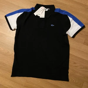 Svart och blå pikétröja från Lacoste - Snygg pikétröja från Lacoste i svart med blå och vita detaljer på axlar och ärmar. Klassisk krage och korta ärmar, med den ikoniska blå krokodilloggan broderad på bröstet. Tröjan är gjord i mjuk bomull och har en normal passform.