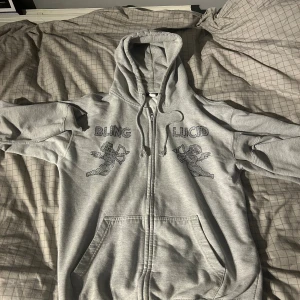Grå hoodie från Blinglucid med änglar - Snygg grå hoodie från Blinglucid med dragkedja och huva. Framsidan har coola svarta ängelmotiv och texten 'BLING' och 'LUCID' i blingad stil. Tröjan har känguruficka och snörning vid huvan. Materialet är 75% bomull och 25% polyester, perfekt för chill dagar.