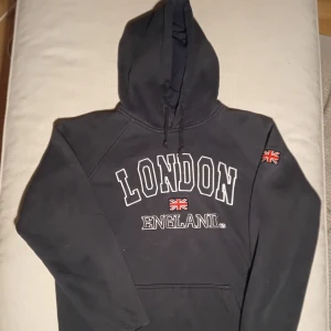 Mörkblå  London hoodie - Svart hoodie med stor vit broderad text 'LONDON ENGLAND' och Union Jack-flagga på bröstet och ärmen. Tröjan har huva med snörning och en klassisk känguruficka framtill. Perfekt för dig som gillar streetwear och vill ha en chill look.
