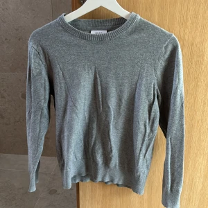 Grå stickad tröja från Grunt - En enkel och stilren grå stickad tröja från Grunt i storlek M. Tröjan har rund hals, ribbade muddar och långärmad passform. Storlek M i barnstorlek 