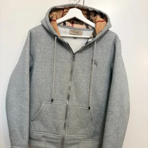 Burberry Zip Hoodie - Snygg grå hoodie från Burberry med dragkedja framtill, snörning i huvan och klassiskt rutigt foder. Broderad logga på bröstet och ribbade muddar. Perfekt för en chill och stilren look. Knappt använt, alla tags finns och kan skickas med!