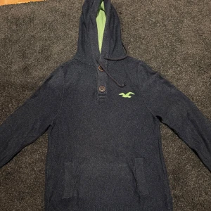 Mörkblå hoodie från Hollister L - Snygg mörkblå hoodie från Hollister med grönt foder i huvan och broderad logga på bröstet. Tröjan har långa ärmar, två bruna knappar framtill och praktisk snörning vid huvan. Perfekt för chill dagar och sköna höstkvällar.