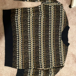 Mönstrad stickad tröja i brunt och blått - Säljer en stickad tröja med snyggt geometriskt mönster i brunt, beige, svart och mörkblått. Tröjan har ribbade muddar vid ärmslut, hals och nederkant. Perfekt för dig som gillar retrovibbar och vill ha något unikt i garderoben.